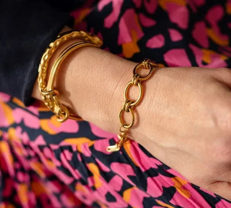 A close-up of a person's wrist wearing a gold chain link bracelet with a small lobster clasp, alongside stacked gold bangles, set against a vibrant pink, orange, and black patterned garment.