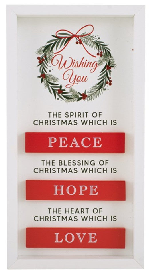 A framed Christmas wall sign with a white frame and a wreath at the top reading 'Wishing You.' Below, black text says 'The Spirit of Christmas which is PEACE,' 'The blessing of Christmas which is HOPE,' and 'The heart of Christmas which is LOVE,' with each word on a red rectangular panel.