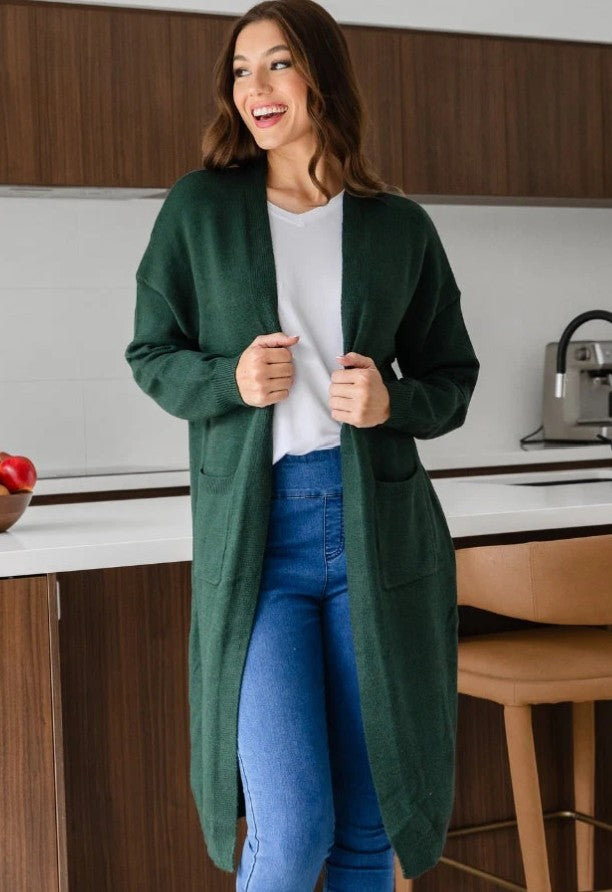 Woman wearing forest green longline knit cardigan over white top and blue jeans in modern kitchen setting.