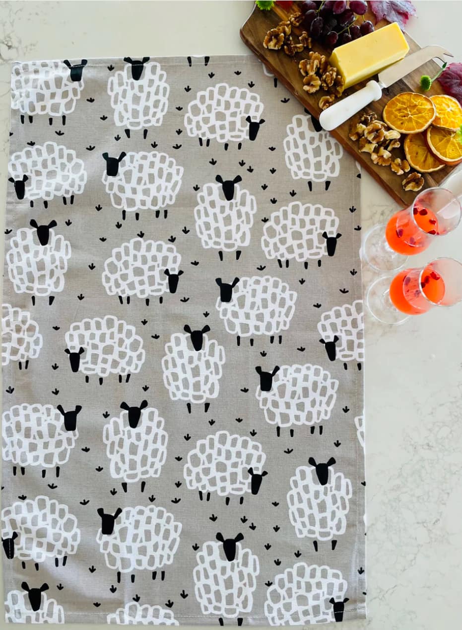 Sheep-patterned tea towel on a table with food and drinks.